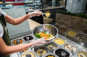 High angle of crop unrecognizable seller in gloves putting assorted ingredients in bowl while working in salad bar in supermarket