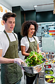 Cheerful diverse sellers in aprons standing at counter in salad bar with fresh food in metal bowls