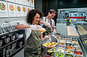 Lächelnde Verkäuferinnen und Verkäufer in Schürzen bereiten Salate in einer Bar in einem modernen Supermarkt zu