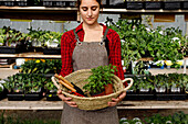 Crop woman in apron carrying wicker basket with gardening tools and potted sprouts while working in hothouse