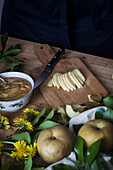 From above pieces of ripe apple and bowl with cream placed on table near dandelions and crop person during pastry preparation in kitchen