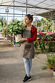 Full body happy woman carrying potted green plant and smiling while working in hothouse