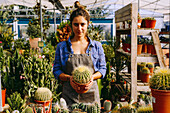 Anonymous person in apron and checkered shirt demonstrating pot with green succulent to camera while working in greenhouse