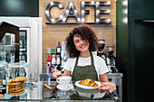 Fröhliche, ethnische Barista-Frau in Schürze steht am Tresen mit einer Tasse aromatischem Cappuccino und einem frischen Croissant und schaut in die Kamera