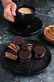 Chocolate alfajores with dulce de leche placed on black plate against blurred background of crop person holding cup