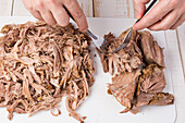 From above crop anonymous woman pulling cooked meat on small pieces and fibers with forks on cutting board while cooking Cuban bowl