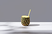 Reife Ananas mit gestreiftem Strohhalm auf einem sonnenbeschienenen Tisch, der ein frisches, gesundes Getränk auf grauem Hintergrund symbolisiert