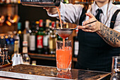 Skilled young female bartender using strainer and pouring red alcohol drink from shaker into glass with ice cubes while preparing refreshing cocktail at bar counter