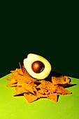 Half of ripe avocado with seed placed on pile of crunchy tortilla chips against dark green background