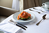 Blick von oben auf einen Teller mit leckerem Avocado-Toast mit pochiertem Ei und geräuchertem Lachs, serviert auf einem Tisch mit Besteck und Kaffeetasse während des Frühstücks im Hotelrestaurant