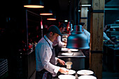 Cook holding spoon and little pan while adding sauce in plate on restaurant kitchen