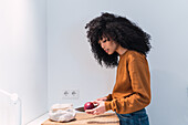 Side view of young black female with curly dark hair cutting fresh apple with knife on kitchen counter and looking down while cooking at home