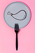 Top view of black plastic fork placed on pink table on gray plate with painted cartoon chicken drumstick