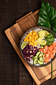 Top view of appetizing colorful poke bowl with fresh salmon and rice served with ripe avocado slices, cabbage and mango placed on wooden tray