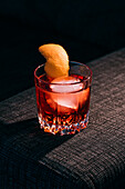 Glass of refreshing Negroni cocktail with bitter flavor and ice garnished with orange peel and served on couch arm in dark room