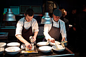 Young cooks men in aprons carefully serving meal in white ceramic dishes on restaurant kitchen
