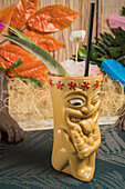 Polynesian tiki cup of cold alcohol beverage decorated with straw and green leaves placed against colorful leaves and dry grass