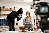 Unrecognizable woman taking photo of chocolate muffins on digital camera against blogger talking during cooking process in kitchen