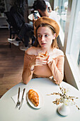 French female in beret sitting at table in cafe with aromatic glass of coffee and freshly baked croissant