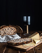 Cut slice of walnut and raisin sourdough bread with loaf on towel served with aromatic cheese and glass of red wine on table