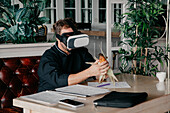 Unrecognizable male chef in uniform sitting at table with various documents and smartphone and holding grilled corn while experiencing virtual reality in VR headset in restaurant
