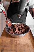 Cropped unrecognizable worker in black apron wearing gloves while melting dark chocolate in stainless steel bowl using heat gun working at counter with marble slab