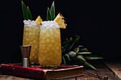 Wooden table with glasses of fresh yellow cocktails with ice cubes and pineapple pieces and leaves near spoon and shot glass placed on red book on black background