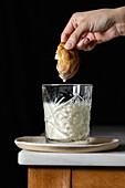 Crop anonymous person dipping fresh sweet cookie into glass of milk placed on corner of table against black background in kitchen