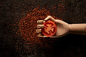 Top view composition with fresh red tomato slice in artificial wooden hand placed above ground sun dried tomatoes on black background