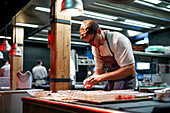 Young serious chef man in cooking apron serving meal in dishes on restaurant kitchen