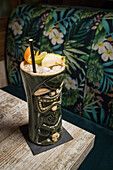 Large sculptural tiki cup filled with booze decorated with straw and fruits placed on green rug against wooden table
