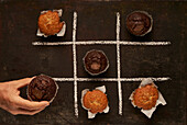 Top view of crop anonymous person putting chocolate muffin in grid while playing Tic tac toe game with desserts on black background