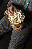 From above cropped unrecognizable kid sitting comfortable on sofa with popcorn bucket while eating and watching TV