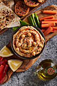 Draufsicht auf eine Schale mit leckerem Hummus, die auf einer Holzplatte mit verschiedenen Gemüsesorten in der Nähe von Zitronenscheiben und einem Krug mit Öl vor Tortilla-Chips und Croutons steht