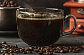 Glass of freshly brewed aromatic coffee on saucer placed on table with scattered beans against manual grinder