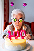 Optimistische ältere Frau mit Spielzeugaugen, die sich amüsiert, während sie an einem Tisch mit einem Kuchen sitzt, der mit Kerzen mit der Zahl 100 verziert ist und ein Jubiläum feiert