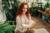 Concentrated red haired female sitting on chair with crossed legs and reading book among different potted plants in domestic garden