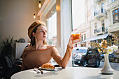 French female in beret sitting at table in cafe with aromatic glass of coffee and freshly baked croissant