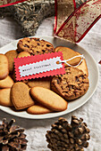 Teller mit verschiedenen süßen Keksen und Geschenkanhänger auf einem Tisch mit Weihnachtsdekoration von oben