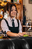 Young woman in uniform chopping fresh lime on counter while preparing cocktail during work in bar