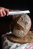 Side view of faceless cook with bread knife cutting whole rye sourdough bread loaf on wooden board against black background