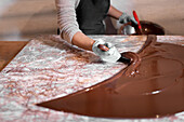 Anonymer Angestellter mit Handschuhen, der mit einem Metallspatel flüssige Schokolade auf dem Tisch verteilt, während er in einer Konditorei ein Dessert zubereitet