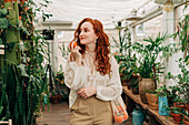 Charming young female with long wavy red hair standing with reusable string bag of tangerines in room with various houseplants