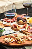 Delicious bacon served on wooden cutting board on table with various appetizers and glasses of red wine