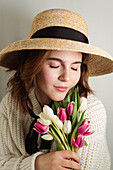 Dreamy female in dress and with delicate tulip flowers standing with closed eyes on white background in studio
