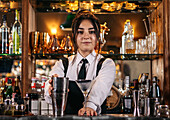 Cheerful female bartender in uniform standing at counter with glass and metal shaker while preparing cocktail and looking at camera