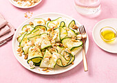 From above fresh salad of thin sliced ripe zucchini, feta cheese and pine nuts in white plate on table pink background