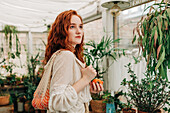 Charming young female with long wavy red hair standing with reusable string bag of tangerines in room with various houseplants