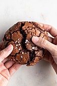 Top view of crop anonymous person breaking freshly baked chocolate brownie cookie with cracks on white background in light kitchen