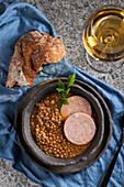 Top view rustic bowl with tasty lentil soup with parsley and slices of sausage placed on marble table and blue napkin near bread and wine during lunch